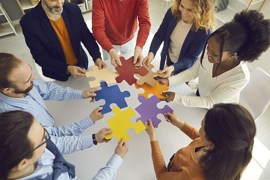 About Our Agency - Portrait of a Diverse Group of Employees Standing in a Conference Room While Holding Colorful Puzzle Pieces
